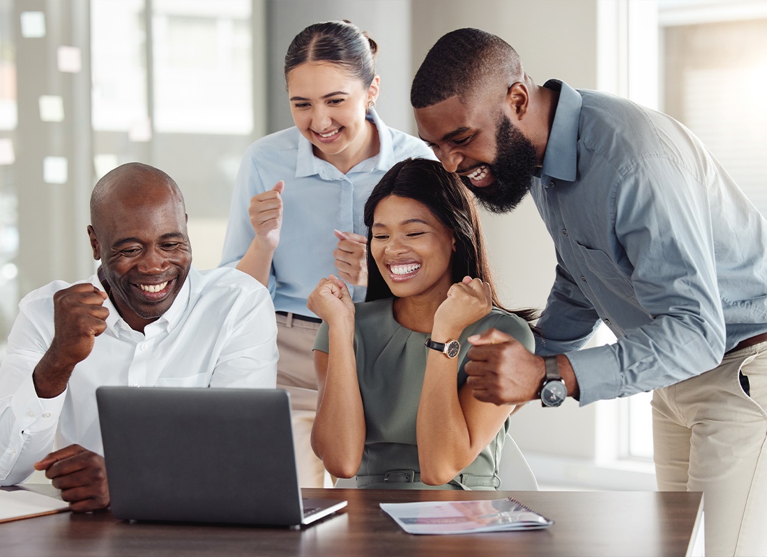 Business Insurance - A Team of Employees Celebrating a Good Job While Sitting and Standing in Front of a Laptop at a Wooden Table